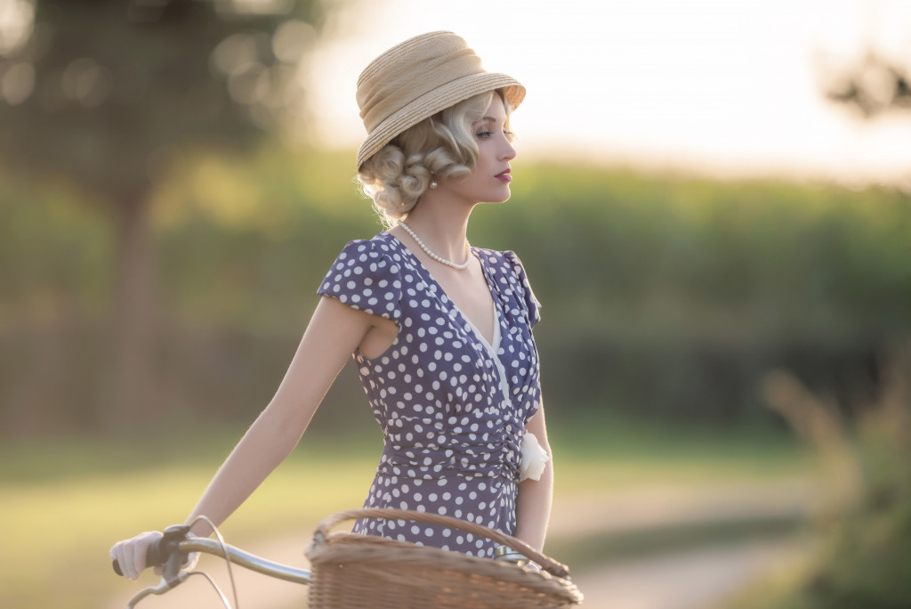 Vintage 1930s fashion woman in blue summer dress standing with bicycle in rural landscape