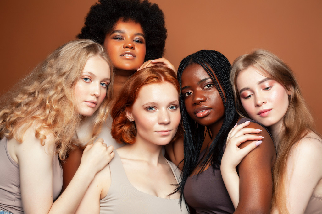 five women of different skin color and type hugging each other in brown background