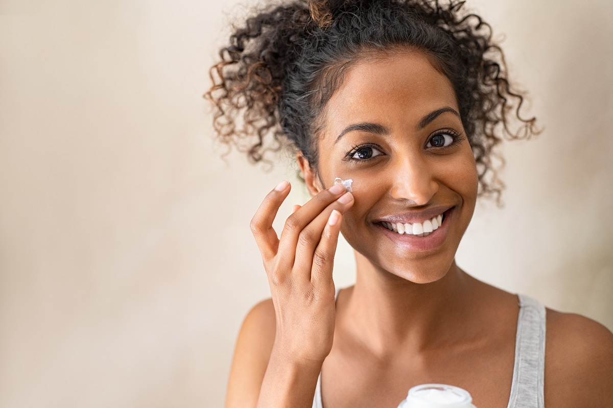 black woman smiling while applying moisturizing cream in her face across a mirror